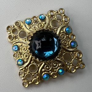 Könnte beinhalten: Eine quadratische, goldfarbene Brosche mit einem komplizierten Filigran-Design aus Metall mit offenen kreisförmigen Mustern. In der Mitte befindet sich ein großer, runder, dunkelblauer Cabochon-Stein, umgeben von acht kleineren, runden, irisierenden hellblauen Strasssteinen.
