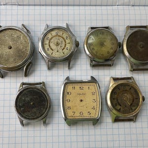 May include: A collection of seven vintage wrist watch cases and dials in various states of wear. The assortment includes round and rectangular metal housings with aged, discolored, or weathered faces, some featuring numerical markers and watch hands.