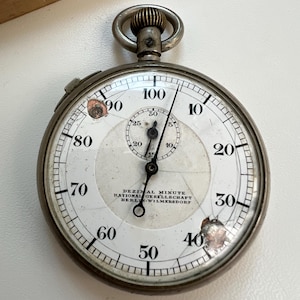 May include: A silver-toned metal stopwatch with a white face, black numerals from 10 to 100, and a smaller sub-dial. The face features the text DEZIMAL MINUTE RATIONAL GESELLSCHAFT BERLIN-WILMERSDORF. The glass cover has visible cracks and damage.