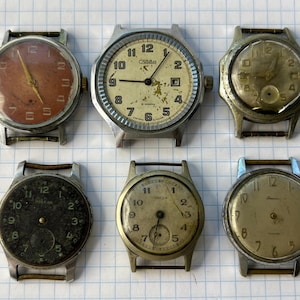 May include: A collection of six vintage wristwatches with round metal cases and various dial colours including copper, cream, black, and silver. Each watch features Arabic numerals, thin hands, and exposed lugs, showing signs of age and wear.
