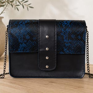 Op de afbeelding: Een rechthoekige schoudertas met een onderkant van zwart leer en een klep met een blauw en zwart slangenleerpatroon. Een verticale zwarte leren band met vier zilverkleurige metalen studs loopt over het midden. Voorzien van een donkere metalen ketting.
