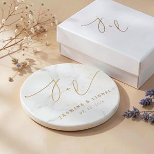 Könnte beinhalten: Ein runder Untersetzer aus weißem Marmor mit goldener Schreibschrift, die J + L, JASMINE & LIONEL und 07. 30. 2026 zeigt, begleitet von einer passenden weißen Geschenkbox mit den goldenen Initialen J + L auf dem Deckel und getrockneten Blumen.