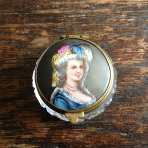 May include: A vintage glass trinket box with a gold metal rim and a painted image of a woman wearing a blue and white feathered hat. The woman is wearing a blue and white dress and a pearl necklace.