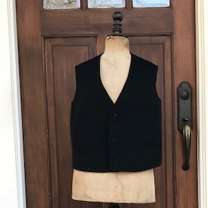 May include: A black wool vest on a vintage wooden dress form with a black base. The dress form is standing in front of a wooden door.