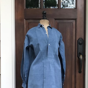 May include: A blue linen shirt with a long, pointed collar and a button-up front. The shirt has a long, flowing hemline and is displayed on a black mannequin.