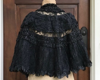 May include: A black lace cape with a ruffled trim. The cape is on a dress form.