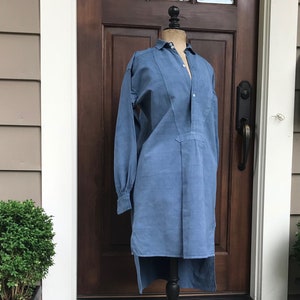 May include: A long-sleeved, light blue shirt with a high collar and a button-up front. The shirt is displayed on a black mannequin stand against a backdrop of a wooden door and a brick wall.