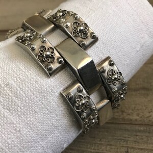 May include: A silver bracelet with a chain link design and intricate floral details. Each link is decorated with a small, raised flower.