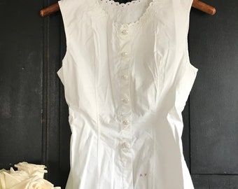 Puede incluir: Blusa blanca sin mangas con cuello de encaje y cierre de botones en la parte delantera. La blusa tiene un corpiño ajustado y un dobladillo ligeramente acampanado.