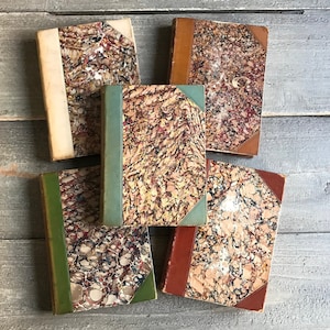 May include: Five antique books with marbled covers in shades of brown, red, green, and white. The books are stacked on a wooden surface.