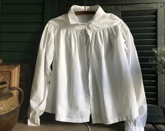 Può includere: Una camicia bianca a maniche lunghe con scollo arricciato e chiusura a cravatta. La camicia è realizzata in un tessuto leggero e ha una vestibilità rilassata.