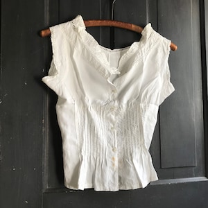 Puede incluir: Una blusa blanca de algodón con cuello de encaje y mangas cortas. La blusa tiene un cierre de botones en la parte delantera y un corpiño ajustado.