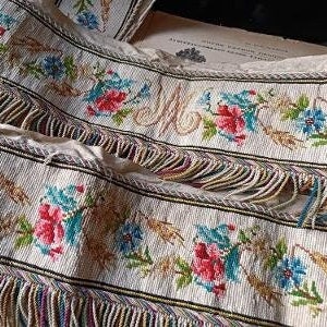 May include: White fabric trim with a floral pattern and fringe. The fabric has a repeating pattern of blue, red, and yellow flowers, and the fringe is made of a variety of colors.