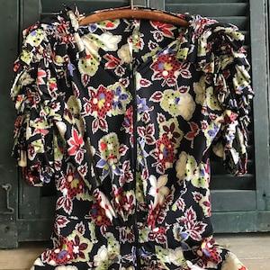 Può includere: Un abito vintage nero con stampa floreale con fiori rossi, verdi e bianchi. L'abito ha un collo e maniche a balze.