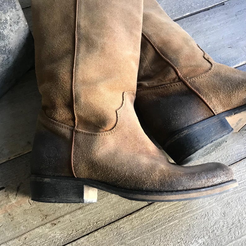 May include: A pair of distressed brown suede knee-high boots. The boots have a worn, vintage appearance with a slightly faded color. They feature a low heel and a classic western-inspired design, perfect for a rustic or bohemian style.