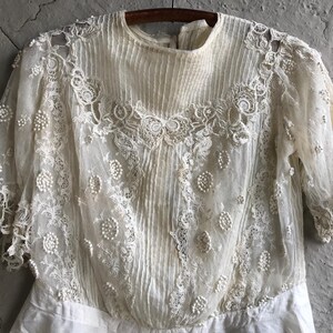 May include: A white lace top with a delicate floral pattern and pearl embellishments. The top has a high neckline and long sleeves.