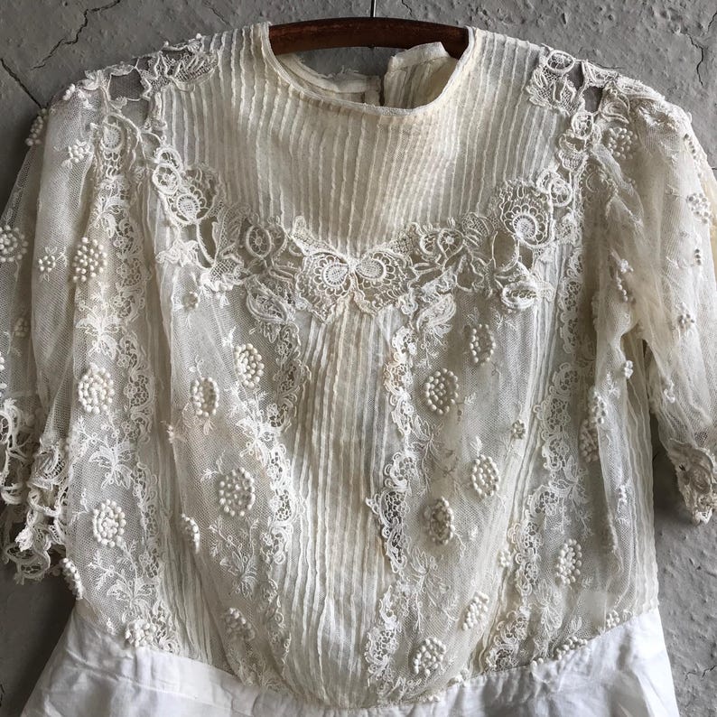 May include: A white lace top with a delicate floral pattern and pearl embellishments. The top has a high neckline and long sleeves.