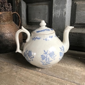 May include: White ceramic teapot with blue floral patterns. The teapot has a curved spout, a curved handle, and a lid with a small finial. The teapot is sitting on a wooden surface.
