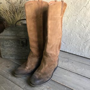 May include: A pair of tall, brown suede boots with a worn, vintage look. The boots have a classic Western style with a pointed toe and a stacked heel. The boots are approximately 16 inches tall.