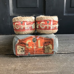Könnte beinhalten: Drei Spulen braunes Garn, verpackt in Papier mit einem roten und weißen Etikett mit der Aufschrift "Comtoir Linier No. 3 Lille Paris Fils de Chanvre Extra 10 Pelotes de 50 Grammes".