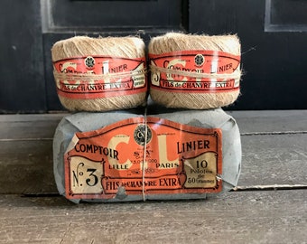 May include: Three spools of brown twine wrapped in paper with a red and white label that reads "Comtoir Linier No. 3 Lille Paris Fils de Chanvre Extra 10 Pelotes de 50 Grammes".