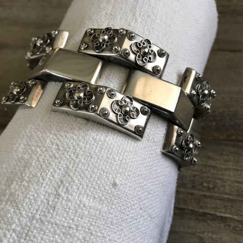 May include: A silver bracelet with a linked chain design. Each link features a decorative floral pattern.