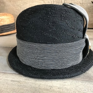 May include: A black straw hat with a wide brim and a black and white striped band. The hat has a small bow on the side.