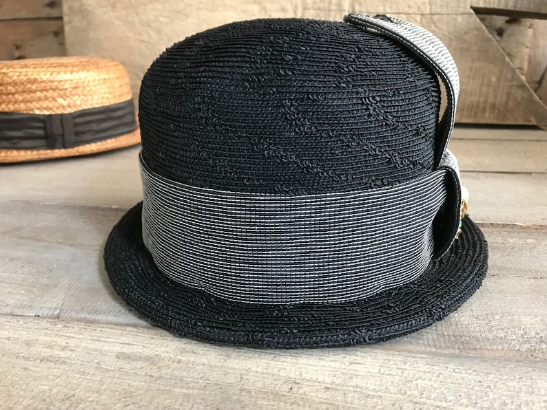 May include: A black straw hat with a wide brim and a black and white striped band. The hat has a small bow on the side.