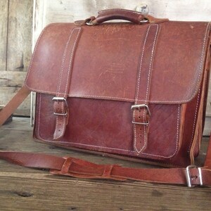 K&ouml;nnte beinhalten: Eine braune Ledertasche mit einem langen Riemen und zwei Schnallen. Die Tasche hat eine Klappe mit einem Griff und einem wei&szlig;en N&auml;hten-Detail.