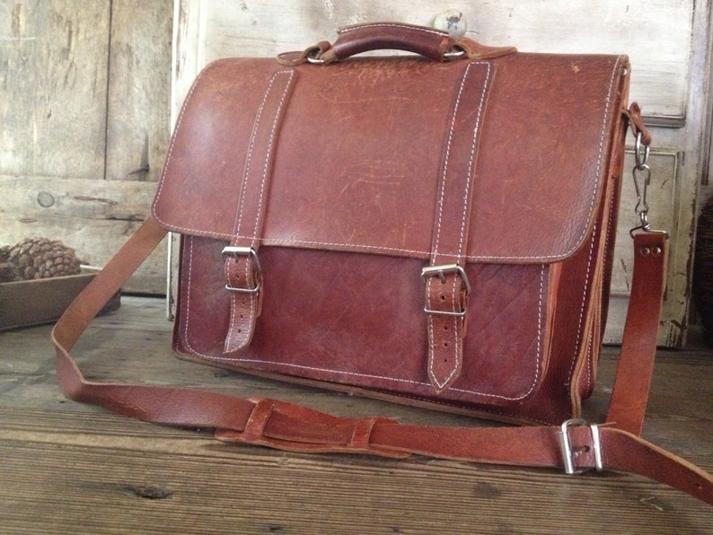 K&ouml;nnte beinhalten: Eine braune Ledertasche mit einem langen Riemen und zwei Schnallen. Die Tasche hat eine Klappe mit einem Griff und einem wei&szlig;en N&auml;hten-Detail.