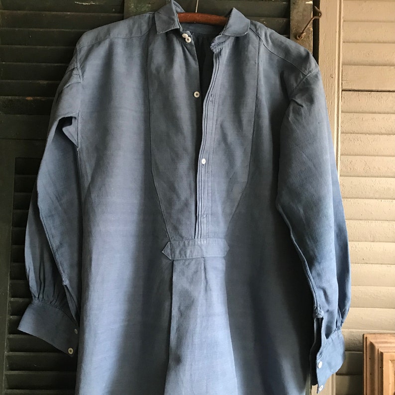 May include: A light blue linen shirt with a button-up front and long sleeves. The shirt has a pointed collar and a single button closure at the neck.