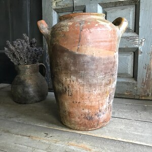 Può includere: Un grande vaso in terracotta usurata con due manici. Il vaso ha una forma cilindrica con un'ampia apertura ed è decorato con sfumature di marrone, arancione e bianco. Accanto c'è un vaso più piccolo, marrone scuro, con lavanda.