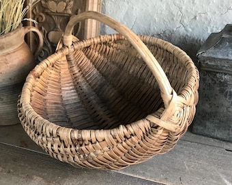 May include: A round, woven basket with a curved handle. The basket is made of light brown wicker and has a natural, rustic appearance. The handle is also made of woven wicker. The basket is empty and sits on a wooden surface.