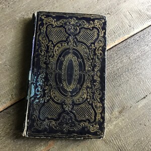 May include: A vintage, black book with gold embossed detailing on the cover. The cover features a decorative design with intricate floral patterns and a central oval.