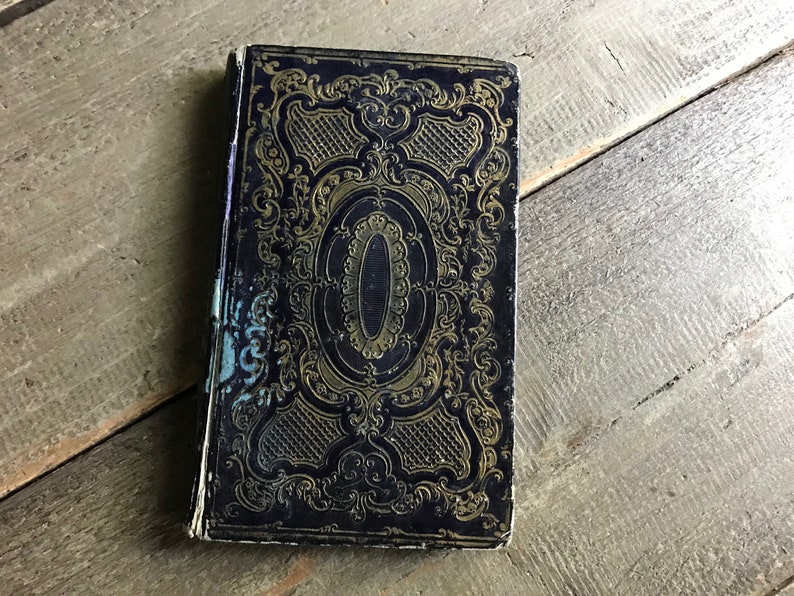 May include: A vintage, black book with gold embossed detailing on the cover. The cover features a decorative design with intricate floral patterns and a central oval.