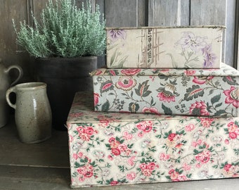 May include: Three rectangular fabric-covered boxes stacked. The boxes have floral patterns in pink, red, and green on a cream background. The top box has a floral and bamboo design. A potted plant and a ceramic jug are in the background.
