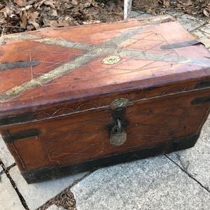 May include: A vintage leather-bound trunk with a brass lock and decorative stitching. The trunk is brown with a light brown and gold design on the top.