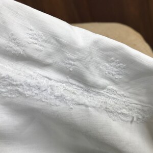 May include: White fabric with a delicate embroidered design. The embroidery features a repeating pattern of small, intricate flowers.