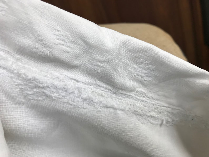 May include: White fabric with a delicate embroidered design. The embroidery features a repeating pattern of small, intricate flowers.