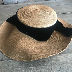 K&ouml;nnte beinhalten: Ein breitkrempiger Strohhut mit einem schwarzen Samtband und Schleife. Der Hut ist hellbraun und hat einen leicht gekr&auml;uselten Rand.