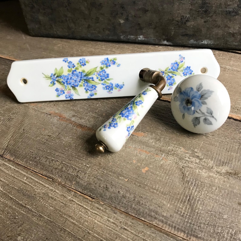 May include: A white porcelain door knob and backplate set with a blue floral design. The knob has a brass stem and the backplate has a small hole for mounting.