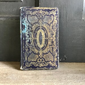 May include: A vintage book with a dark blue cover and gold ornate design. The cover features a central oval with a gold border and intricate floral patterns.