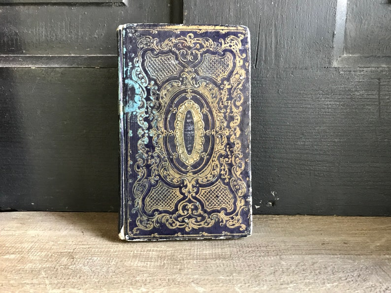 May include: A vintage book with a dark blue cover and gold ornate design. The cover features a central oval with a gold border and intricate floral patterns.