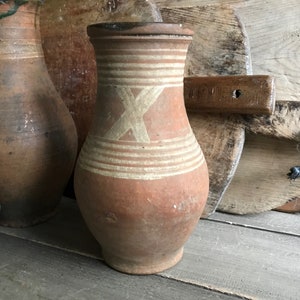 May include: Two brown clay jugs with white painted designs. The jug on the right has a white X design and white stripes. The jugs are sitting on a wooden surface.