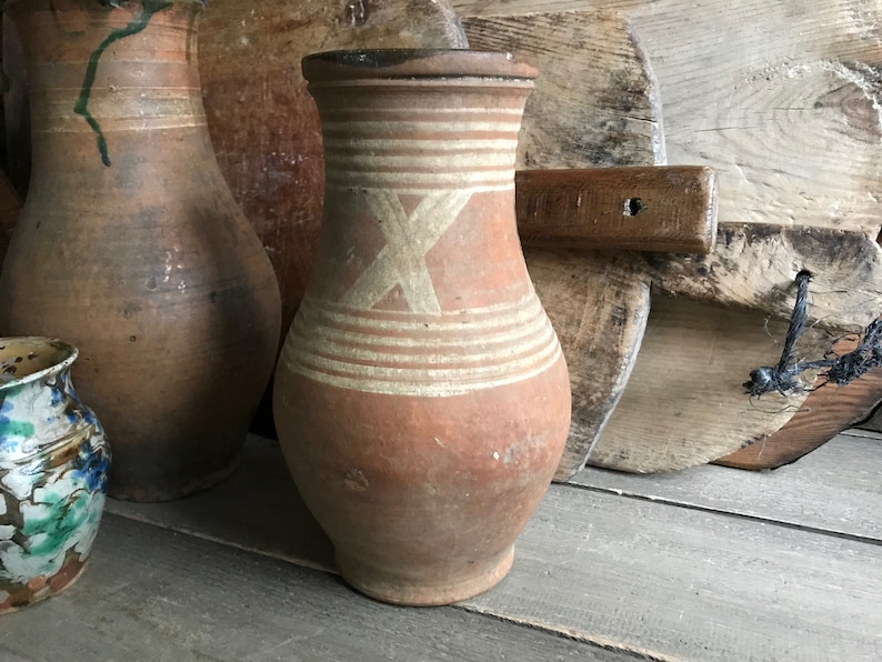 May include: Two brown clay jugs with white painted designs. The jug on the right has a white X design and white stripes. The jugs are sitting on a wooden surface.