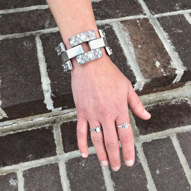May include: A silver bracelet with a geometric design and a row of small rhinestones. The bracelet is worn on a person's wrist. The person is also wearing two silver rings on their fingers.