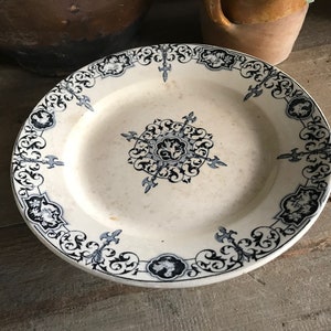 Puede incluir: Un plato de cerámica blanco con un diseño floral azul y blanco. El plato tiene un borde festoneado y un centro elevado.