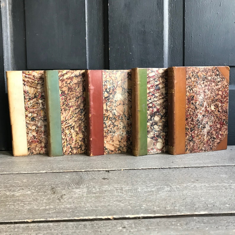 May include: Five antique books with marbled covers and different colored spines. The books are arranged in a row on a wooden surface.