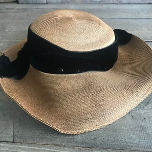 K&ouml;nnte beinhalten: Ein breitkrempiger Strohhut mit einem schwarzen Samtband und Schleife. Der Hut ist hellbraun und hat eine leicht gebogene Krempe.