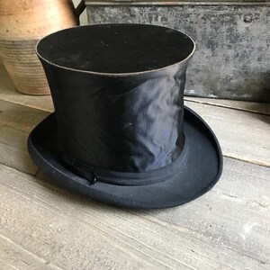 May include: A black top hat with a satin finish. The hat has a tall, cylindrical crown and a wide, flat brim.